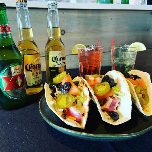 Three tacos filled with toppings, accompanied by two bottles of Corona and two cocktails on a black table.