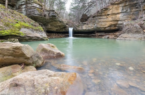 A serene landscape featuring a small waterfall cascading into a clear pool, surrounded by rocky terrain and lush greenery.