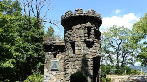 Stone tower with a castle-like design, surrounded by trees and blue sky, featuring a plaque on the front.