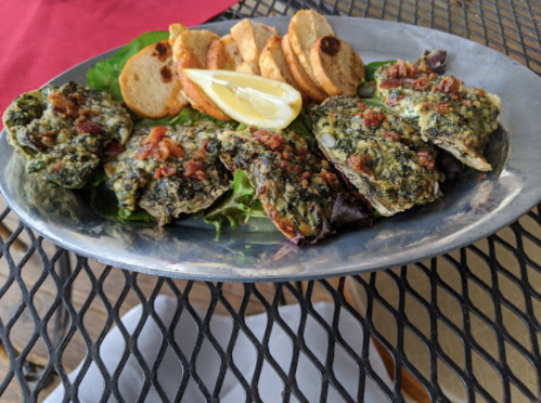 A platter of baked oysters topped with herbs and bacon, served with lemon wedges and toasted bread slices.