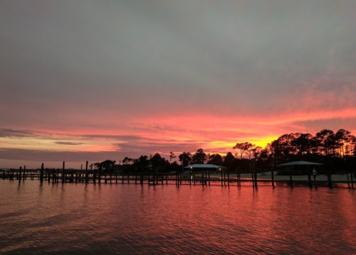 A vibrant sunset over calm waters, reflecting shades of orange and pink, with silhouettes of trees and a pier in the foreground.