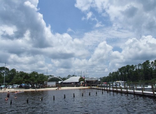 A scenic view of a beach with people swimming, surrounded by trees and boats under a cloudy sky.
