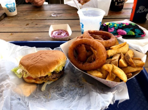 A burger with lettuce and tomato next to a serving of onion rings and fries on a wooden table.