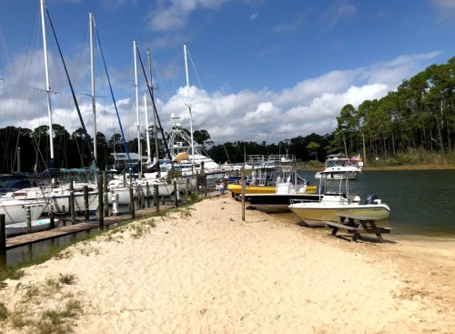 A sandy shore lined with boats and sailboats, surrounded by trees under a partly cloudy sky.