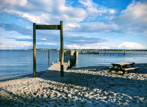 A sandy beach with a wooden pier extending into the water, framed by clouds and blue sky. A picnic table is nearby.