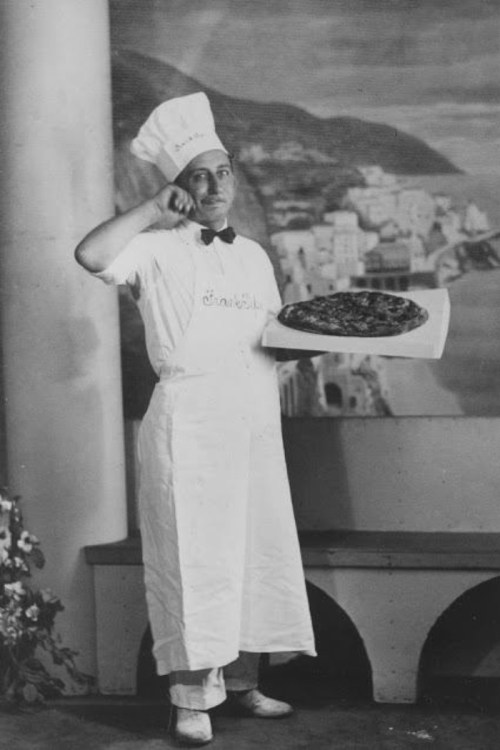A vintage black-and-white photo of a chef holding a large pizza, wearing a chef's hat and apron, with a scenic backdrop.