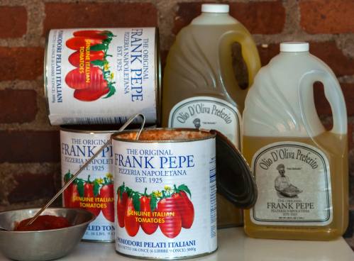 Cans of tomatoes and bottles of olive oil from Frank Pepe Pizzeria, set against a brick background.