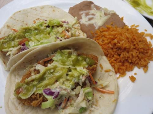 Two tacos filled with fried fish and slaw, served with rice and refried beans on a white plate.
