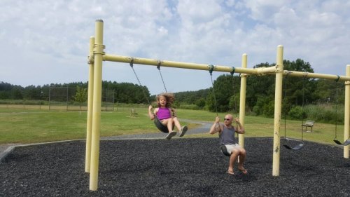 A child swings joyfully on a playground while an adult sits nearby, enjoying a sunny day outdoors.