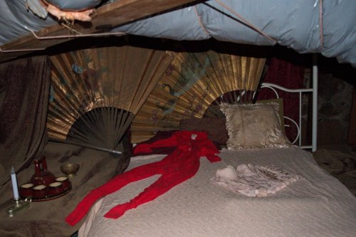 A dimly lit room with a bed covered in a quilt, featuring a red outfit and decorative fans in the background.