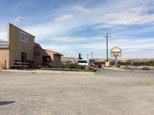 A roadside café with a sign, surrounded by gravel and sparse landscape under a blue sky.