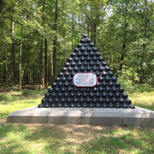 A pyramid-shaped monument made of black balls, surrounded by trees, with a plaque at the front.