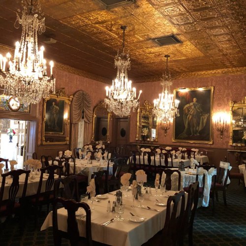 Elegant dining room with chandeliers, ornate decor, and tables set for a formal meal. Richly detailed walls and artwork.
