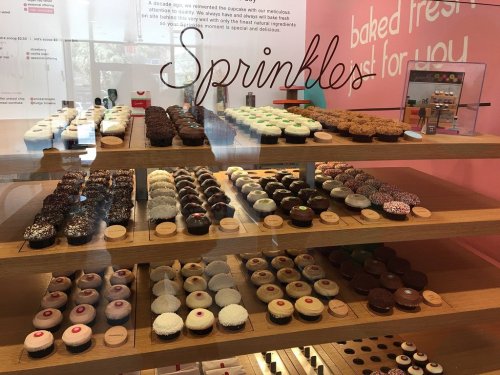 A display case filled with a variety of colorful cupcakes and desserts, labeled "Sprinkles" in a bakery setting.