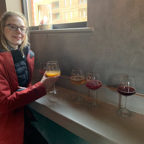A person in a red jacket sits at a table with five colorful drinks in glasses, smiling at the camera.