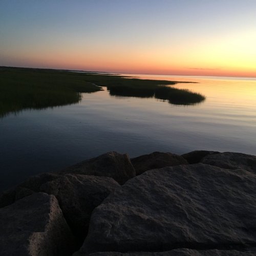 A serene sunset over calm waters, with grassy marshes and rocky foreground, creating a peaceful coastal scene.