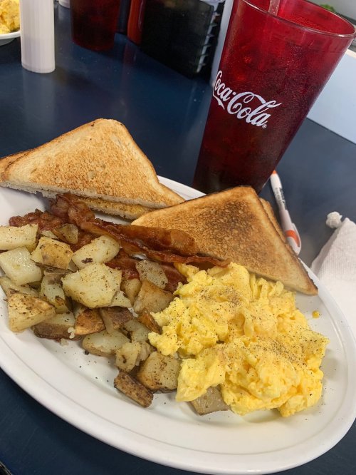 A plate of breakfast featuring toast, bacon, scrambled eggs, and seasoned potatoes, with a Coca-Cola glass in the background.