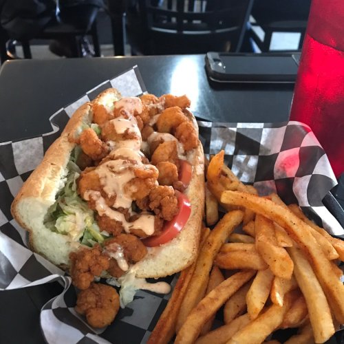 A shrimp po' boy sandwich with lettuce and tomatoes, drizzled with sauce, served with a side of fries and a drink.