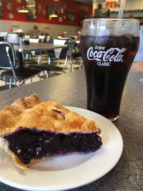 A slice of blueberry pie on a plate next to a glass of Coca-Cola in a retro diner setting.