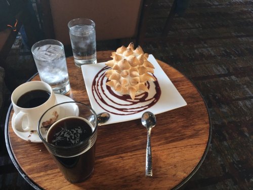 A wooden table with a dessert topped with meringue, chocolate sauce, and several glasses of water and coffee.