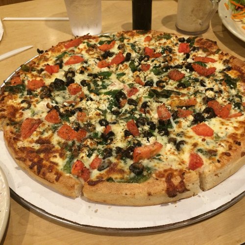 A freshly baked pizza topped with tomatoes, olives, spinach, and cheese, served on a round platter.