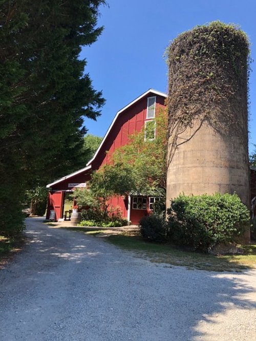 A red barn with a cylindrical silo, surrounded by trees and a gravel path on a sunny day.