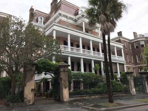 Historic brick mansion with white columns, surrounded by greenery and palm trees, in a charming urban setting.