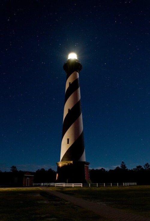 A striped lighthouse illuminated at night, surrounded by a starry sky and a dark landscape.