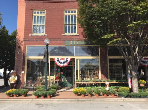A charming storefront with large windows, patriotic decorations, and potted plants lining the entrance.
