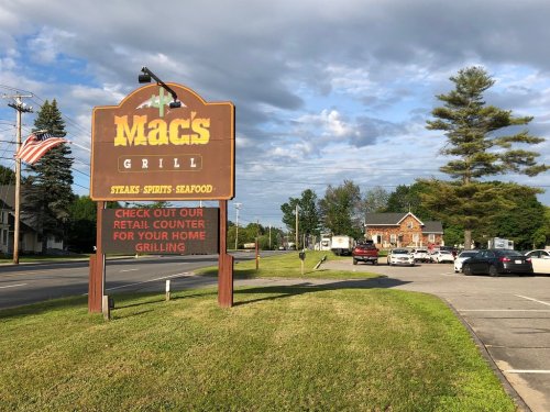 Sign for Mac's Grill, featuring steaks, spirits, and seafood, with a grassy area and parked cars in the background.