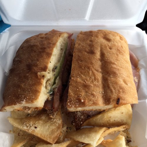 A sandwich with layers of meat and vegetables, served with a side of crispy chips in a takeout container.