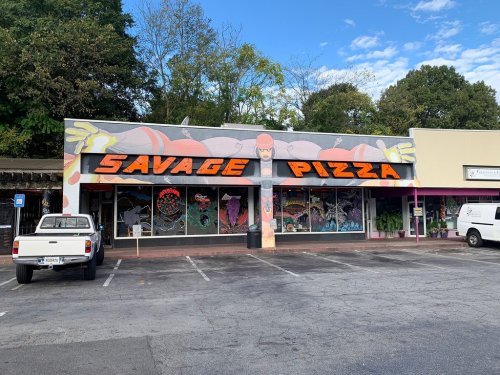 A colorful storefront of "Savage Pizza" featuring bold lettering and vibrant window art, surrounded by parked cars.