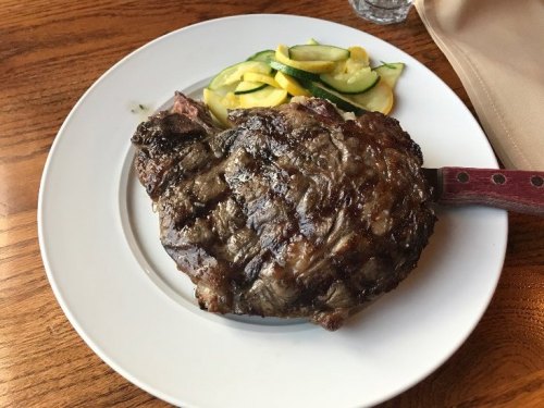 A grilled ribeye steak on a white plate, accompanied by sautéed zucchini and yellow squash.
