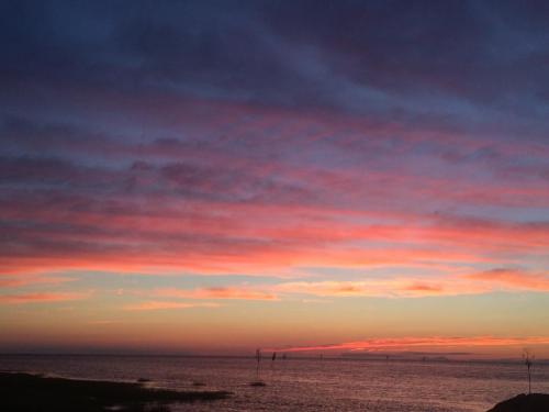 A vibrant sunset over the water, with pink and orange clouds reflecting in the calm sea. Silhouettes of structures in the distance.
