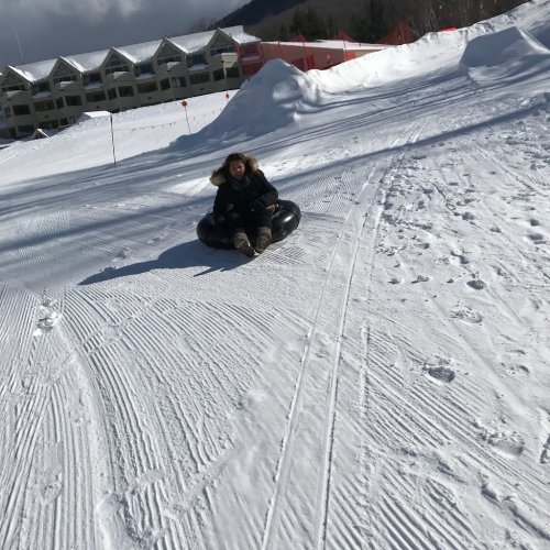 A person in winter clothing sits on a black inner tube, sliding down a snowy slope with buildings in the background.