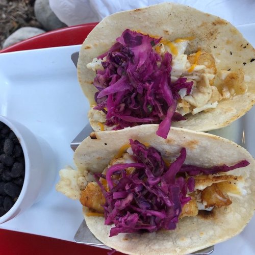 Two tacos filled with fish, cheese, and topped with purple cabbage, served with a side of black beans.