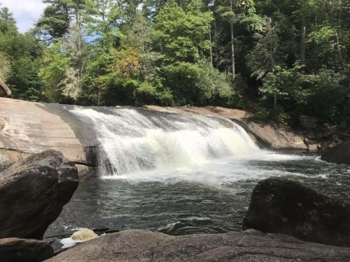A serene waterfall cascades over smooth rocks, surrounded by lush green trees and a clear blue sky.