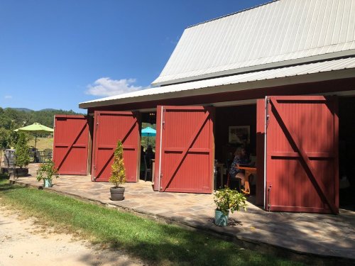 A red barn with open doors, surrounded by greenery and outdoor seating under umbrellas on a sunny day.