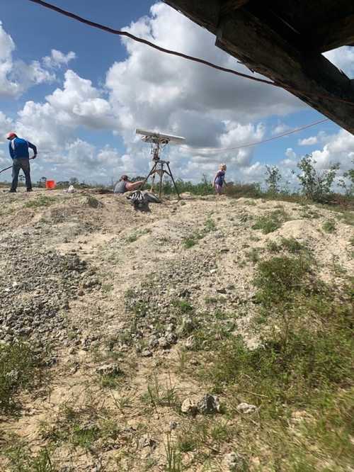 A group of people working outdoors near a drone on a sandy area with scattered vegetation and a blue sky.