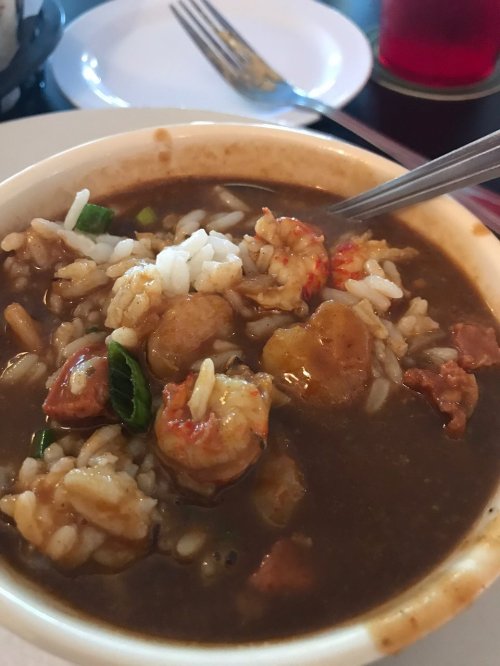 A bowl of gumbo with shrimp, sausage, and rice, garnished with green onions, on a table with a fork and plate in the background.