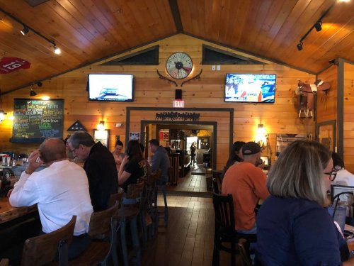 Interior of a rustic restaurant with wooden decor, patrons at the bar, and TVs showing sports.