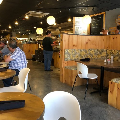 A cozy café interior with wooden tables, a person at the counter, and patrons seated in a booth. Soft lighting and decor visible.