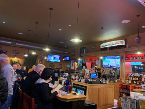 A lively bar scene with patrons at the counter, multiple TVs showing sports, and a well-stocked bar in warm lighting.