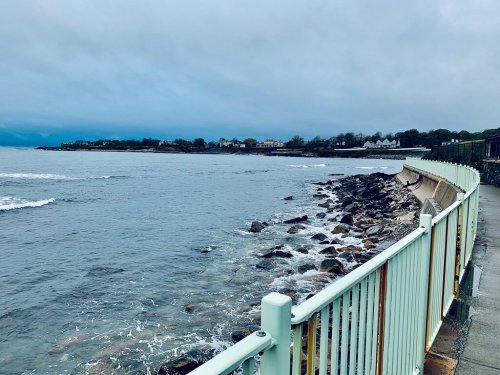 A coastal view featuring a rocky shoreline, gentle waves, and a walkway with a light green railing under a cloudy sky.