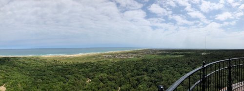 Panoramic view of a coastal landscape with green vegetation, sandy beach, and a blue sky with scattered clouds.
