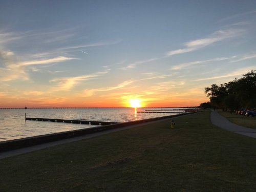 Sunset over a calm lake, with a pier extending into the water and a grassy path along the shore.