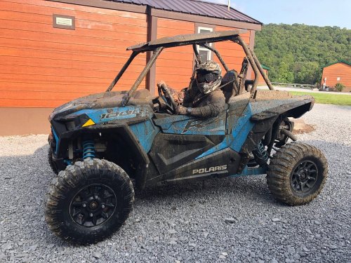 A muddy off-road vehicle parked on gravel, with a driver in a helmet sitting inside, surrounded by trees and buildings.