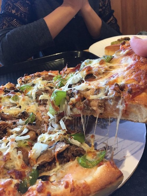 A hand pulling a slice of cheesy pizza topped with meat and green peppers, with a person’s hands clasped in the background.