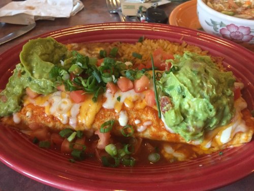A colorful plate featuring a cheesy enchilada topped with guacamole, diced tomatoes, and green onions, served with rice.