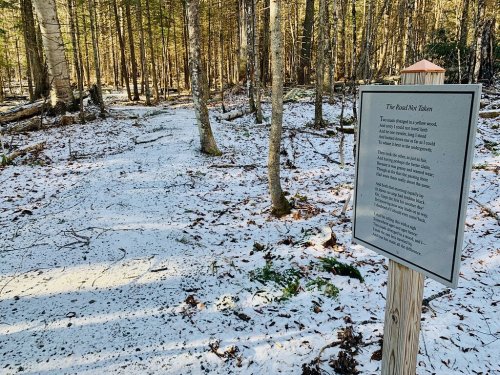 A snowy forest path with a sign displaying a poem titled "The Road Not Taken" among trees.